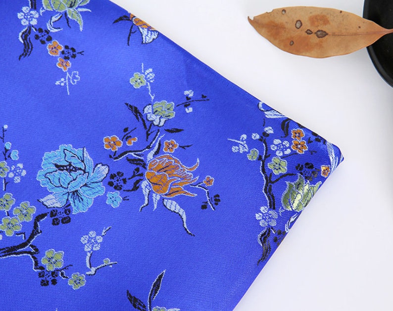 May include: Blue fabric with a floral pattern. The fabric features a repeating design of flowers and leaves in shades of blue, green, orange, and white.