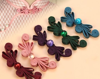 Op de afbeelding: Vijf decoratieve stoffen knopen in verschillende kleuren, waaronder roze, paars, groen, blauw en bordeauxrood. Elke knoop heeft een parelmoerachtig midden en een uniek, ingewikkeld ontwerp.