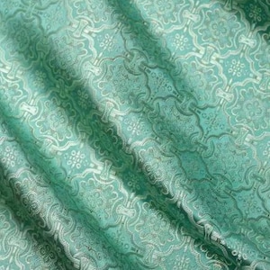 May include: A light mint green fabric featuring a repeating geometric pattern with intricate floral and quatrefoil motifs woven in a lighter, shimmering thread. The material has a soft, draped texture with visible folds creating light and shadow.