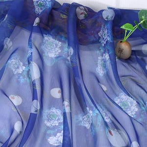 May include: A sheer, flowing fabric in shades of blue, adorned with a delicate floral pattern featuring white and light blue peonies. The fabric is draped, creating soft folds and shadows. A small vase with a plant is visible.