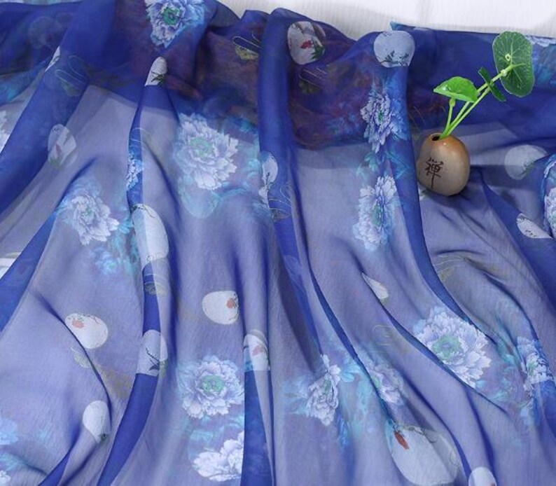 May include: A sheer, flowing fabric in shades of blue, adorned with a delicate floral pattern featuring white and light blue peonies. The fabric is draped, creating soft folds and shadows. A small vase with a plant is visible.