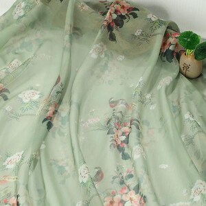 May include: A light green fabric with a floral pattern featuring pink and white flowers, birds, and foliage. The fabric is draped, showing its texture and design. A small vase with greenery is visible in the background.