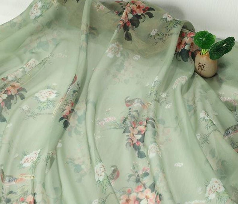 May include: A light green fabric with a floral pattern featuring pink and white flowers, birds, and foliage. The fabric is draped, showing its texture and design. A small vase with greenery is visible in the background.