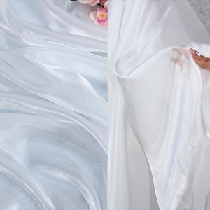 May include: Close-up of white sheer fabric with a soft, flowing texture. The fabric is slightly translucent, allowing light to pass through, creating a delicate and airy appearance. The folds and drapes of the fabric add depth and visual interest.