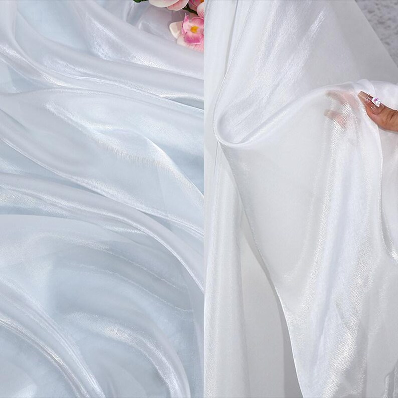 May include: Close-up of white sheer fabric with a soft, flowing texture. The fabric is slightly translucent, allowing light to pass through, creating a delicate and airy appearance. The folds and drapes of the fabric add depth and visual interest.