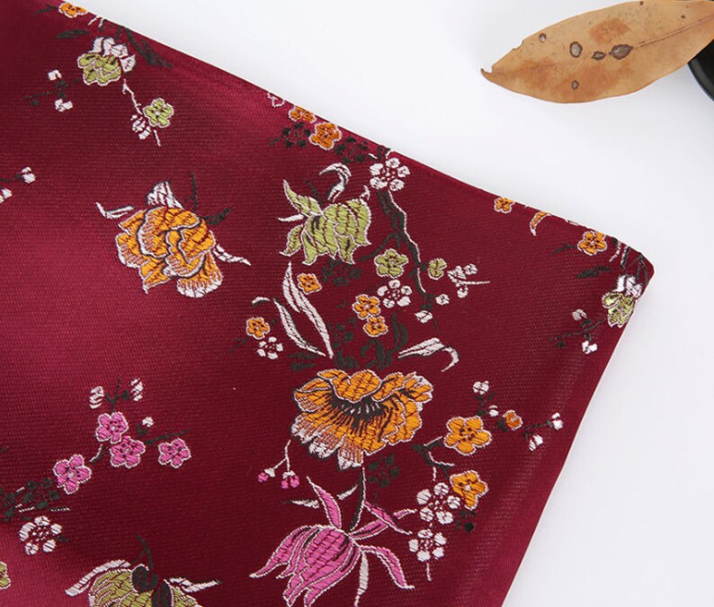 May include: Burgundy fabric with a floral pattern featuring pink, yellow, and green flowers. The fabric has a subtle sheen and a textured surface.