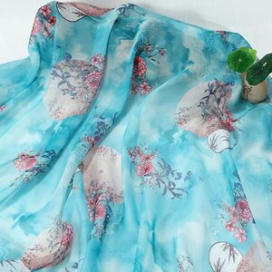 May include: A sheer, light blue fabric with a watercolor-like design. The fabric features large floral patterns in shades of pink and red, with accents of white and green. The overall effect is soft and flowing, suitable for apparel or home decor.