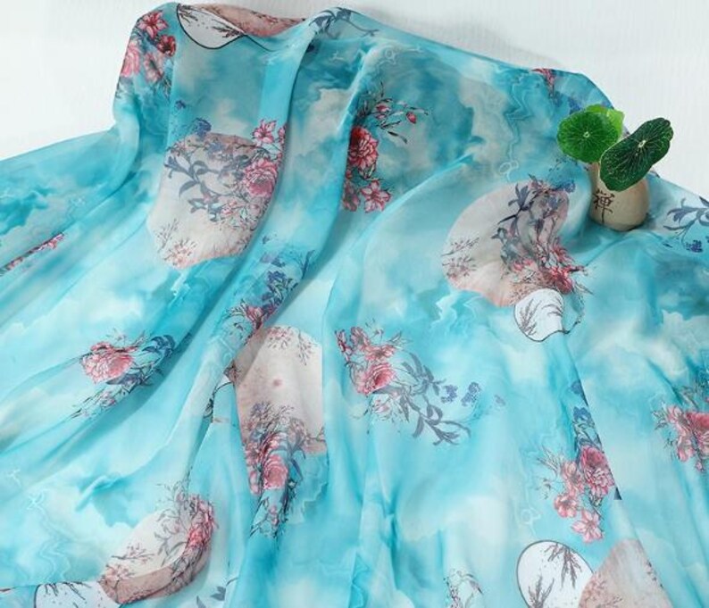 May include: A sheer, light blue fabric with a watercolor-like design. The fabric features large floral patterns in shades of pink and red, with accents of white and green. The overall effect is soft and flowing, suitable for apparel or home decor.