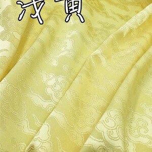 May include: A light yellow fabric featuring a repeating woven pattern of stylized clouds. The material has a subtle sheen and includes the Chinese characters 浅黄 printed in black with a white outline at the top of the textile surface.