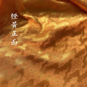 May include: A lustrous orange fabric featuring a repeating woven pattern of stylized clouds. The material has a smooth, reflective surface with deep folds. The Chinese characters 橙黃正面 are printed vertically in white on the left side.