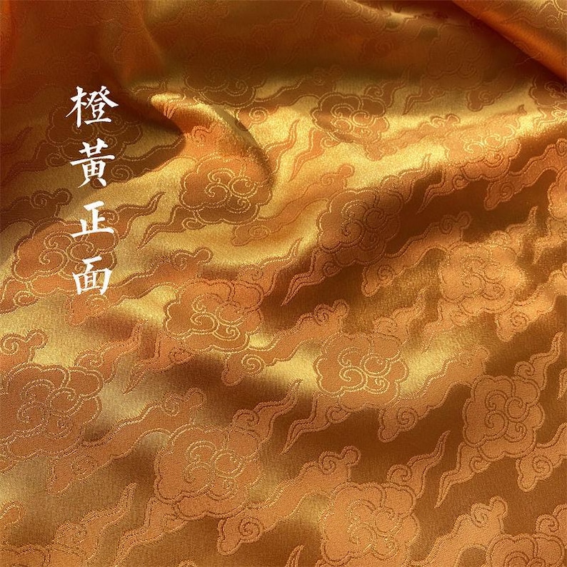 May include: A lustrous orange fabric featuring a repeating woven pattern of stylized clouds. The material has a smooth, reflective surface with deep folds. The Chinese characters 橙黃正面 are printed vertically in white on the left side.
