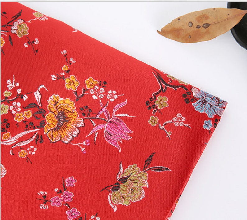May include: Red fabric with a floral pattern. The flowers are in shades of pink, yellow, and blue. The fabric has a subtle sheen.