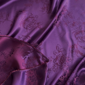 Può includere: Un tessuto lussuoso, viola intenso con un motivo floreale. Il tessuto è drappeggiato, mostrando pieghe e ombre che evidenziano l'intricato design. Il motivo floreale è di una tonalità di viola più scura.