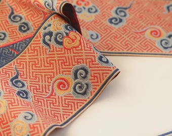 May include: A long fabric strip featuring a repeating geometric fretwork pattern in orange and red. The textile is decorated with embroidered cloud motifs in shades of blue, yellow, and red, with a dark blue border along the edges of the material.