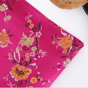 May include: A close-up of a pink fabric with a floral pattern. The fabric features a repeating design of orange, yellow, and green flowers on a pink background.
