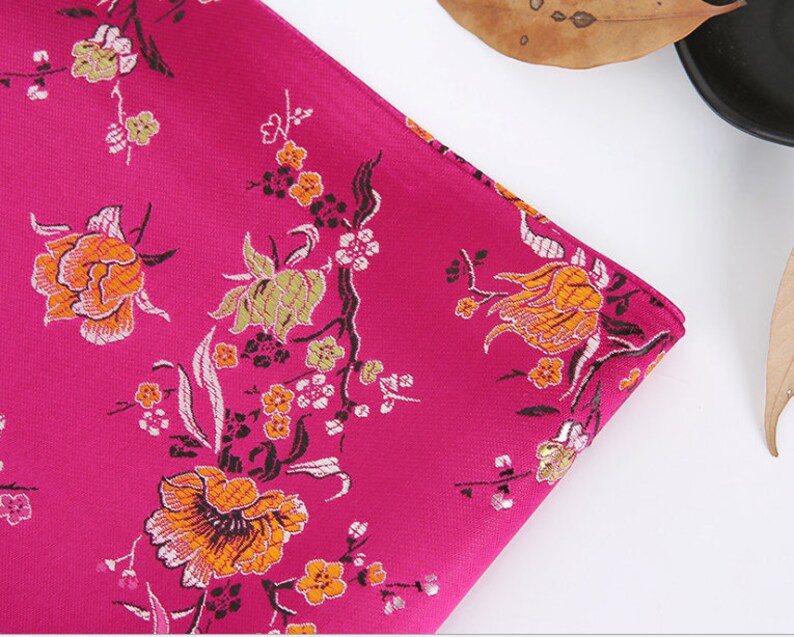 May include: A close-up of a pink fabric with a floral pattern. The fabric features a repeating design of orange, yellow, and green flowers on a pink background.