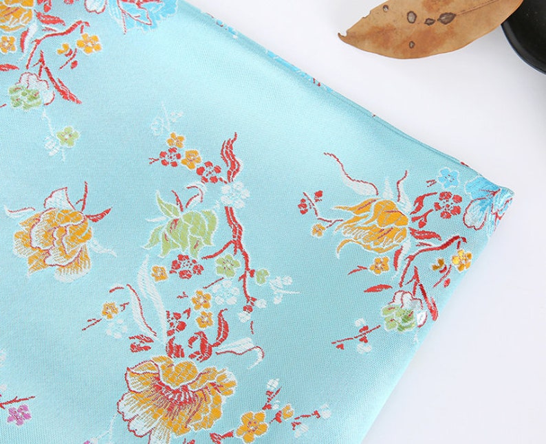 May include: A light blue fabric with a floral pattern. The flowers are yellow, orange, red, and white. The fabric has a subtle sheen.