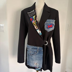 May include: A black blazer featuring a comic book print on the left lapel, a denim pocket with a red and blue BANG patch, and a large denim panel on the lower right side. The garment includes a waist belt secured with a silver metal ring.