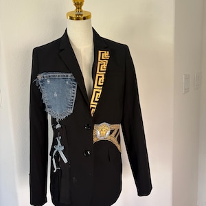 May include: A black blazer featuring a gold Greek key pattern on the left lapel, a blue denim pocket patch on the right chest, and a gold Medusa head emblem on a patterned waist band. The garment has two black buttons and frayed denim accents.