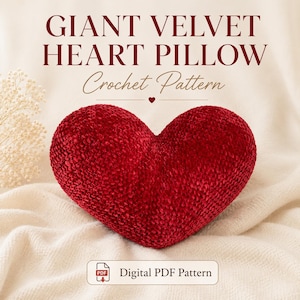 May include: A large, heart-shaped pillow made of red velvet. The pillow is the focal point, resting on a cream-colored fabric. Text at the top reads "GIANT VELVET HEART PILLOW Crochet Pattern." A small icon indicates a digital PDF pattern.