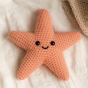 May include: A peach-colored, crocheted starfish plush toy with a smiling face. The starfish has five points and black button eyes. The toy is made of a soft, textured yarn and is suitable for children.
