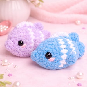 May include: Two handmade crochet fish toys. One is light purple with white accents, the other is light blue with white accents. Both have black eyes and pink cheeks. The background is a soft pink with pearls and flowers.