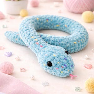 Puede incluir: Un juguete de serpiente de ganchillo azul claro con ojos negros y una lengua rosa. La serpiente está hecha de hilo suave y moteado y está rodeada de elementos decorativos como pompones rosas, cuentas en forma de corazón y una madeja de hilo rosa. Un juguete hecho a mano lindo y tierno.
