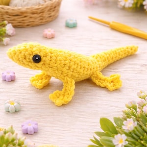 May include: A handmade, yellow crochet lizard with a black eye, lying on a light wood surface. The lizard is surrounded by small flower-shaped decorations and a crochet hook. A basket with yarn balls is in the background.