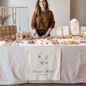 May include: A jewelry display featuring necklaces, earrings, and bracelets. The jewelry is presented on wooden stands and display cards. A banner with the text "Samira Hadid Jewelry store" is draped over the table. The jewelry is gold and silver in color.