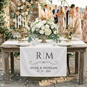 Puede incluir: Una mesa de madera rústica preparada para una boda. Tiene un camino de mesa blanco con las iniciales "RM", los nombres "Rosa & Morgan" y la fecha "27.07.2028". La mesa está decorada con flores, velas y cristalería.