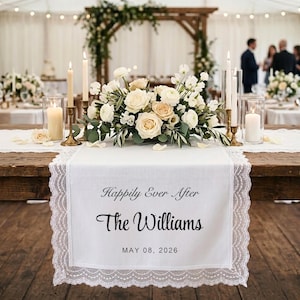 May include: A white table runner with lace edging, inscribed with "Happily Ever After" and "The Williams" with a date. The runner is adorned with a floral arrangement and candles, set for a wedding reception.