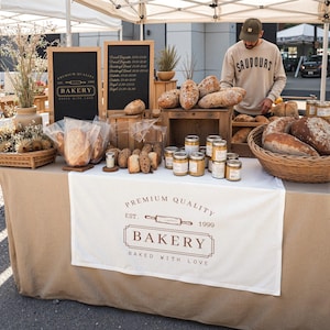 May include: A bakery stand displays various breads, including baguettes and loaves, alongside jars of preserves and baked goods. A sign reads "Premium Quality Bakery, Baked with Love." The stand is covered with a tan cloth and a white banner with the bakery logo.