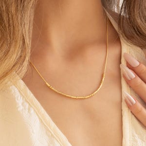 May include: A delicate gold necklace with a minimalist design. The necklace features a thin chain with small, evenly spaced cylindrical beads. The jewelry is worn against a cream-colored top, creating a subtle contrast. The overall aesthetic is elegant and understated.