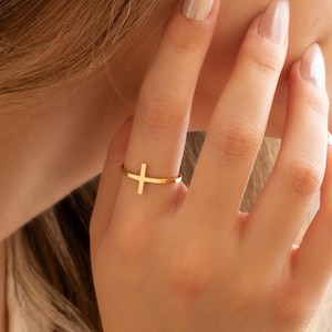 May include: A gold-colored ring featuring a simple cross design. The ring is worn on a finger, with the cross oriented horizontally. The ring appears to be made of a shiny metal, reflecting light.