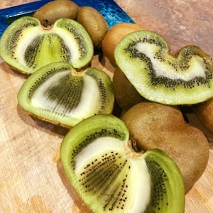May include: A close-up of several kiwis, some whole and some halved. The halved kiwis reveal a vibrant green flesh with black seeds and a white core. The skin is brown and fuzzy. The kiwis are on a wooden surface.
