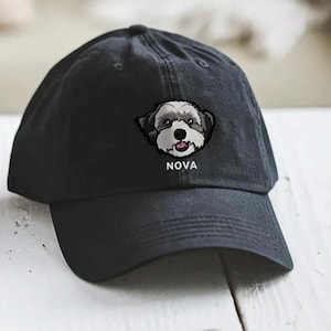 Puede incluir: Gorra de béisbol negra con visera curva. La gorra presenta un parche bordado con la cara de un perro en negro, blanco y gris, con el nombre "NOVA" bordado debajo. La gorra está hecha de una tela suave y duradera.
