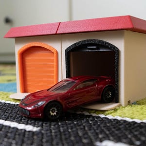 May include: A red toy sports car exiting a toy garage. The garage has a red roof, an orange door, and a black-trimmed garage door. The car is on a black and white road-like surface.
