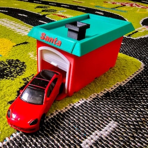 May include: A red toy car exiting a red garage with a green roof. The garage has the word "Santa" printed on it. The toy is on a colorful play mat with a road design.