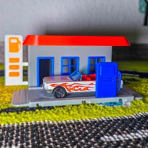 May include: A toy gas station with a red roof, gray walls, and blue doors and windows. A white convertible with orange flames is parked at the gas pump. The gas pump is blue and the car has black wheels.