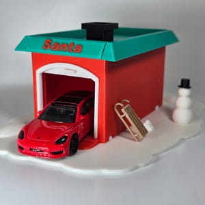 May include: A red toy car is parked inside a red garage with a green roof that reads "Santa". A small snowman and a wooden sled are also present on a white, snow-like base. The scene is festive and holiday-themed.