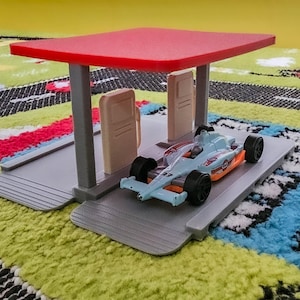 May include: A toy race car in light blue and orange sits on a gray platform under a red canopy. The toy car has black wheels and the number 23 on its side. The scene is set on a colorful rug.
