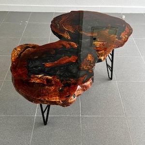 May include: A distinctive coffee table composed of two sections with a live edge. The table showcases a deep brown wood grain with black resin detailing. It is supported by black metal hairpin legs, providing a contemporary contrast to the natural wood.
