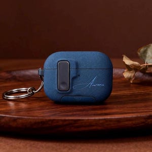 May include: A navy blue AirPods case with a silver keyring. The case features a rectangular button and the cursive word "Aurora". The case is on a wooden tray.