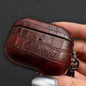 May include: A brown, textured case for wireless earbuds, featuring a crocodile skin pattern. The case has the name "Arlonia" engraved on the front and a silver-colored clasp. The case is held in a person's hand.