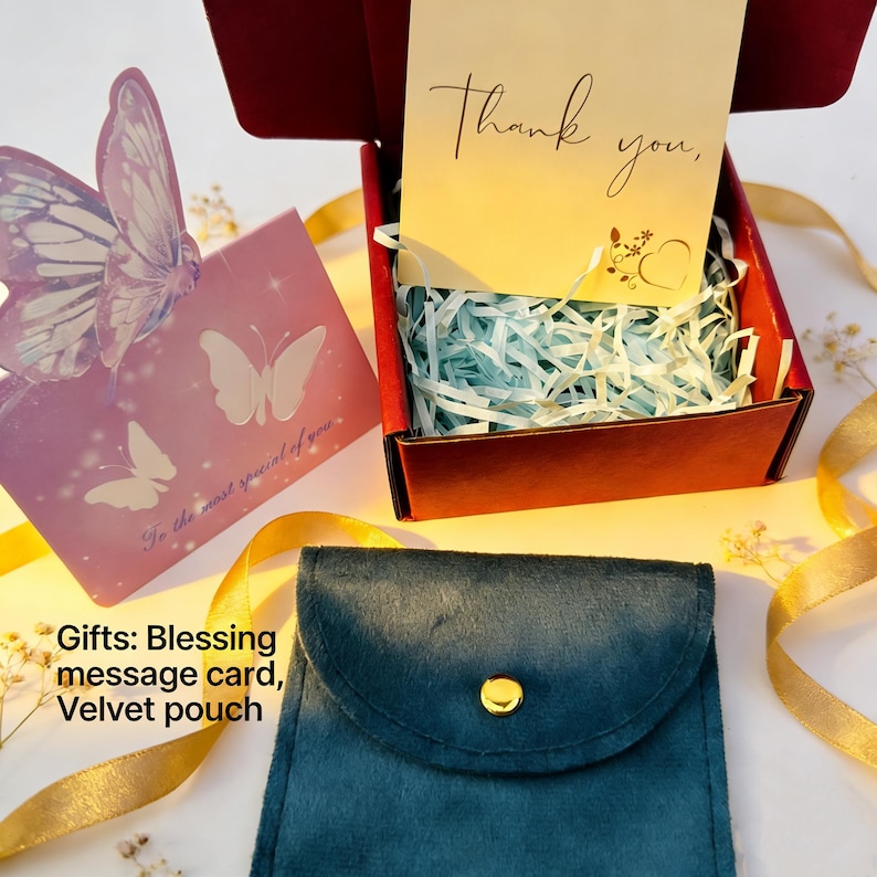 May include: A teal velvet pouch with a gold button, a red gift box with a "Thank you" card, and a pink butterfly card. The gift box contains blue shredded paper. A gold ribbon and small white flowers are also present.