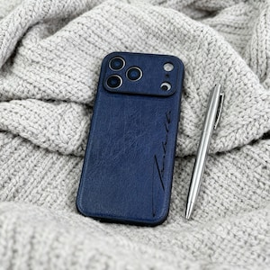 May include: A navy blue phone case with a textured leather appearance, featuring a camera cutout and a stylized signature. A silver pen rests beside the case on a grey knitted fabric.
