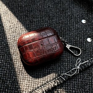 May include: A brown leather AirPod case with a crocodile skin texture. The case has a silver clasp and the name "Antonia" is inscribed on the front. The case is resting on a dark gray textured fabric.