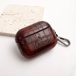 May include: A brown leather AirPod case with a crocodile skin texture. The case has the name "Antonia" engraved on the front and a silver-colored carabiner clip attached to the side.
