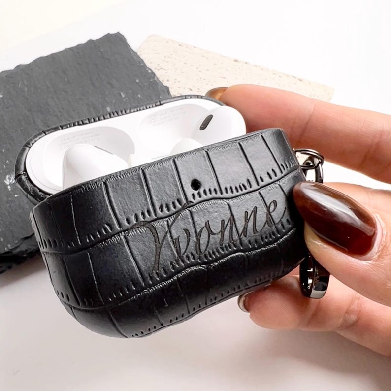 May include: Black leather AirPod case with a crocodile skin texture. The case is open, revealing white AirPod earbuds. The name "Yvonne" is inscribed on the case. A silver-colored clasp is attached to the case.