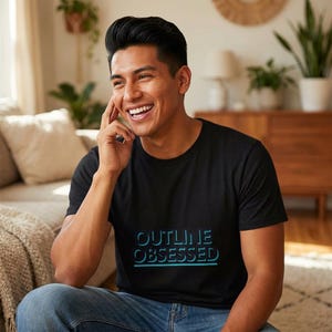 Puede incluir: Camiseta negra de cuello redondo con el texto "OUTLINE OBSESSED" en degradado azul. La persona sonríe y lleva vaqueros azules.